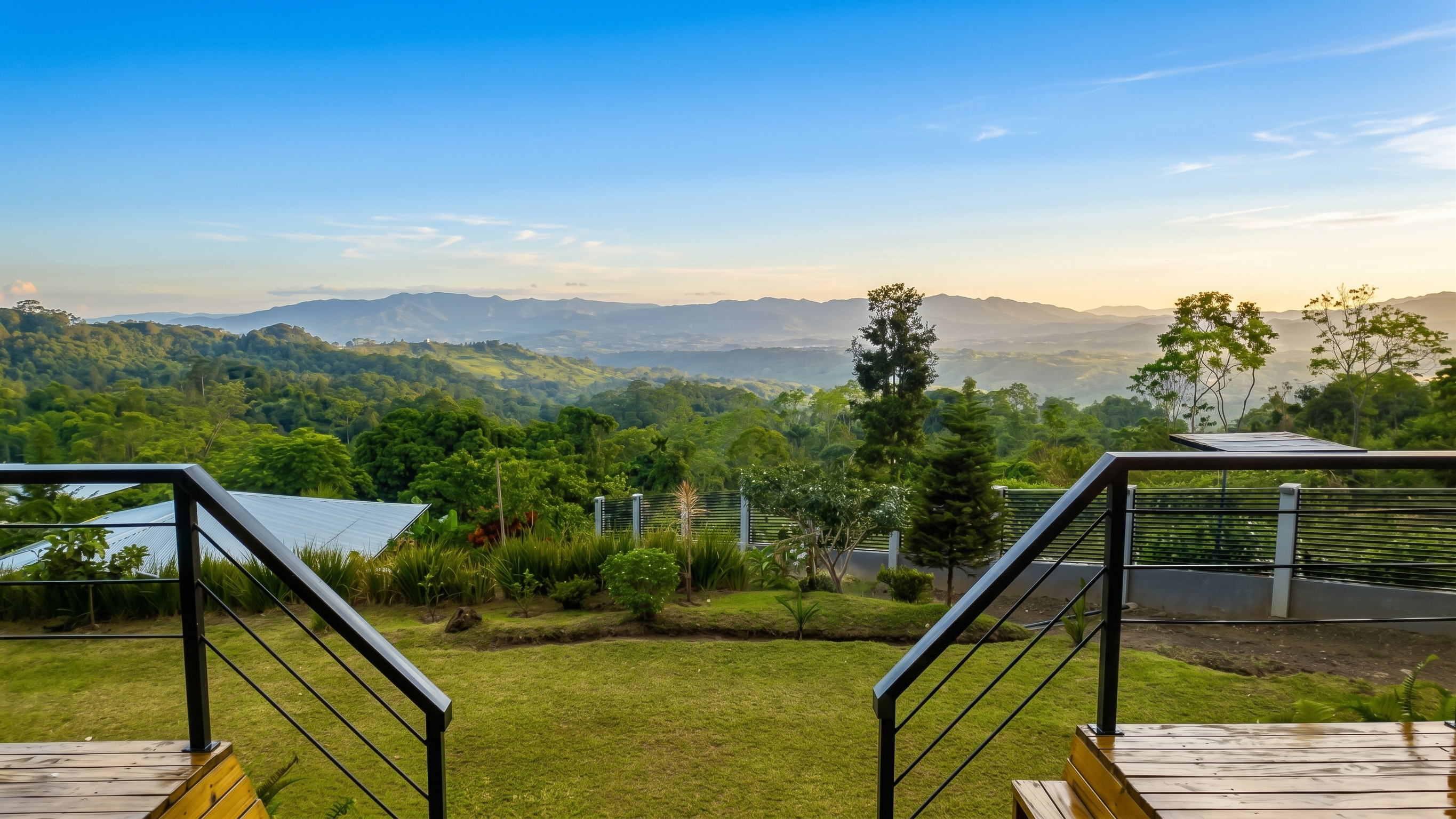 View from a wooden deck across a green garden and rolling hills toward distant mountains, beneath a blue sky with warm light along the horizon.