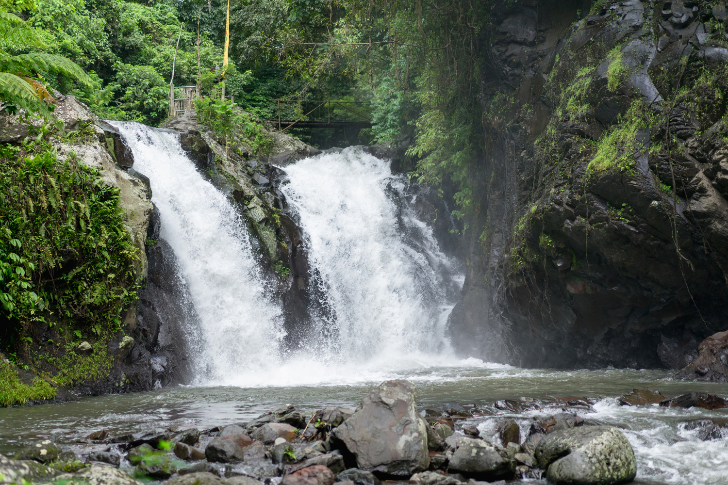 Waterfall cascading over dark rocks into a rocky river, surrounded by lush tropical forest.