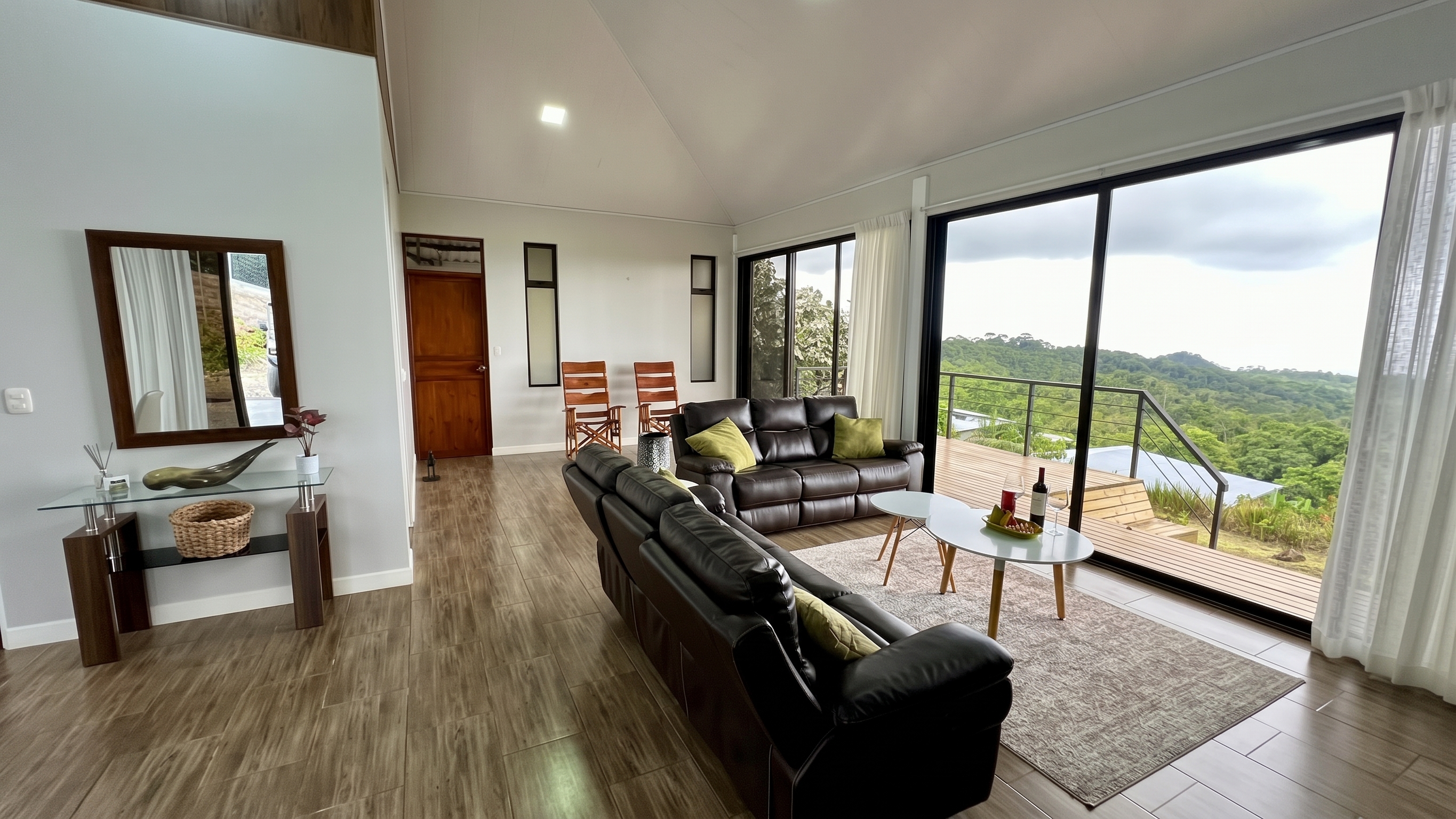 Bright living room with leather sofas, green cushions, and large sliding glass doors opening onto a wooden deck overlooking lush Costa Rican hills.
